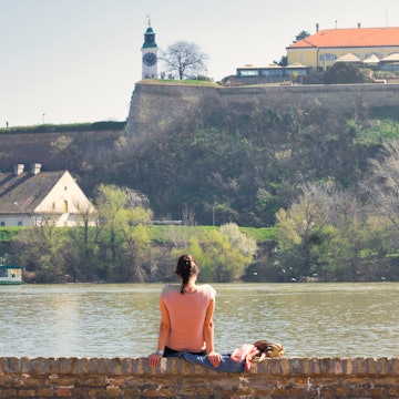 A young woman sitting on the banks of the Danube river in Novi Sad, Serbia, and enjoying the view of the old Petrovaradin fortress; Shutterstock ID 1348024769; your: Claire Naylor; gl: 65050; netsuite: Online Ed; full: Novi Sad weekend update